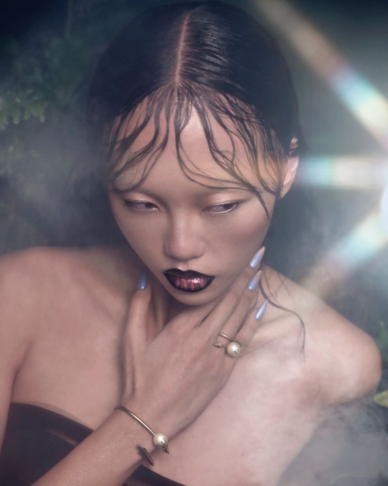 Portrait of a woman with wet-look hair, dark lipstick, and jewelry. The photo features soft lighting and a nature-inspired background with visible fog.