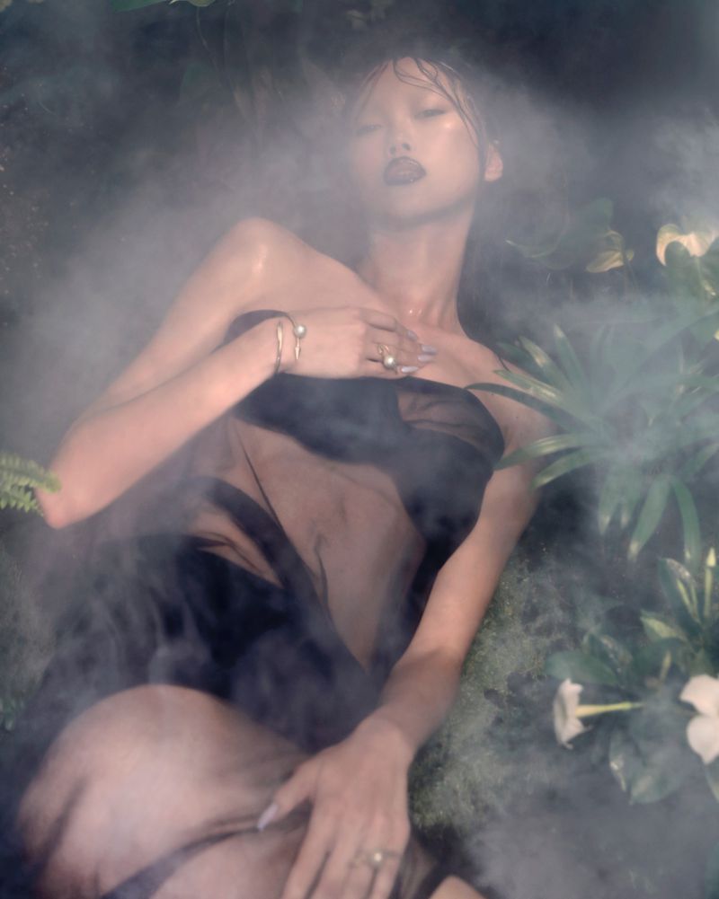 Woman in sheer black fabric reclines among white flowers and green foliage, surrounded by fog. Studio shot with dark, muted tones.