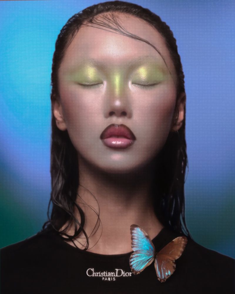 Close-up portrait of a woman with closed eyes and bright eye makeup wearing a black shirt with butterfly pin and 'Christian Dior' logo.