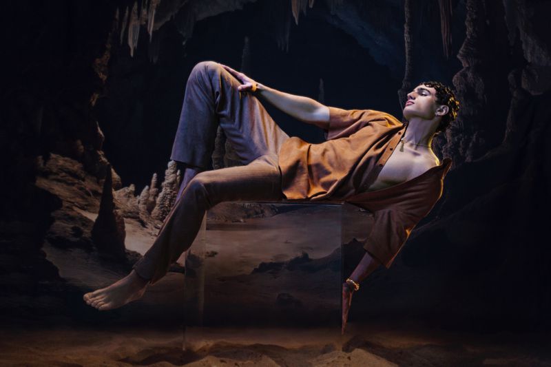 Adult male model poses on a transparent cube in a dimly lit cave with rock formations and earthy colors, showcasing fashion and artistry.