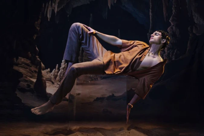 Adult male model poses on a transparent cube in a dimly lit cave with rock formations and earthy colors, showcasing fashion and artistry.