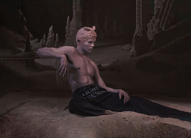 Man with pale skin and blonde hair is reclining on a textured surface in a dark, cave-like setting, wearing jeans with text detail.