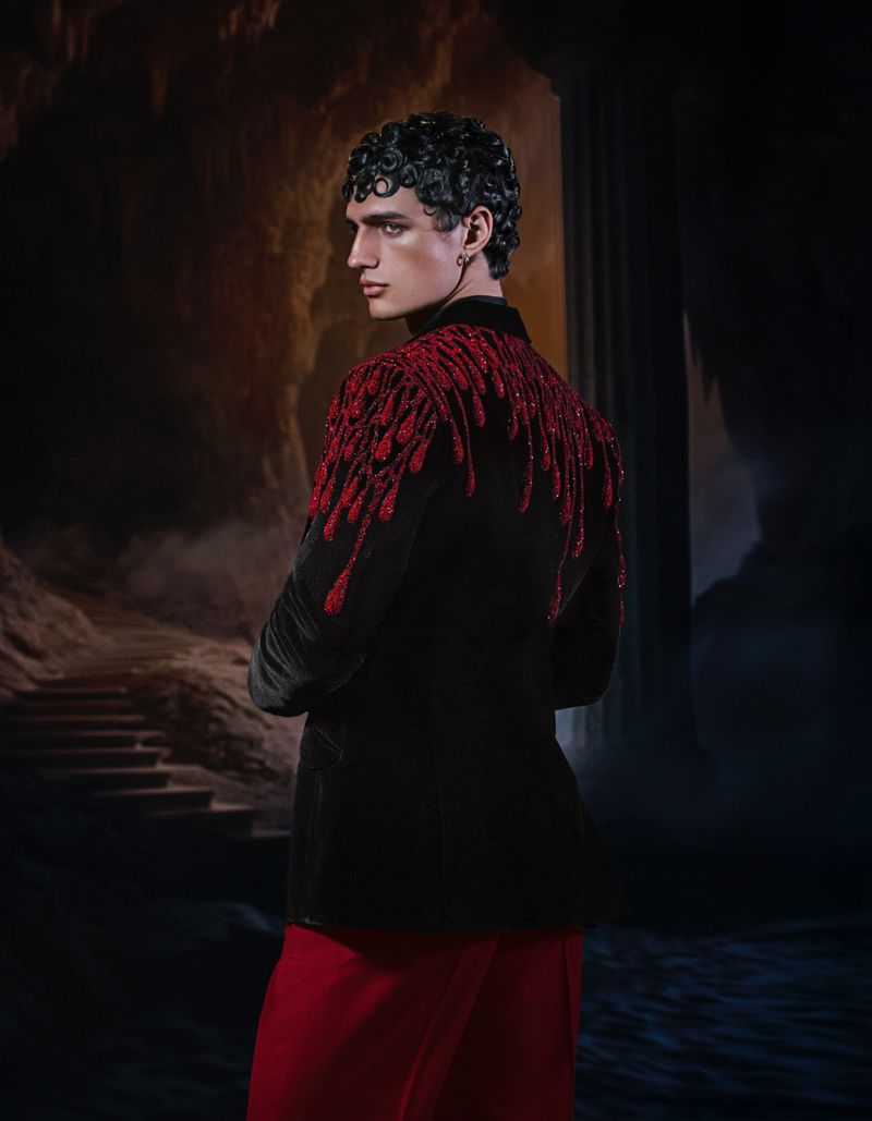 A fashion shot of a man with dark curly hair wearing a black velvet jacket with red sequin embellishments, and red pants against a dark backdrop.