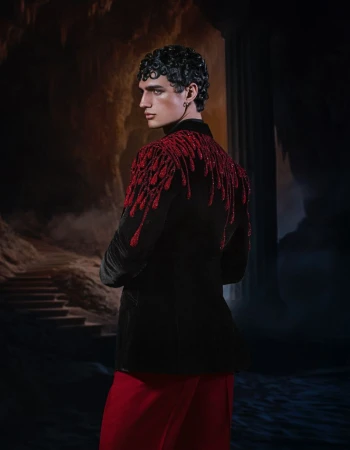A fashion shot of a man with dark curly hair wearing a black velvet jacket with red sequin embellishments, and red pants against a dark backdrop.