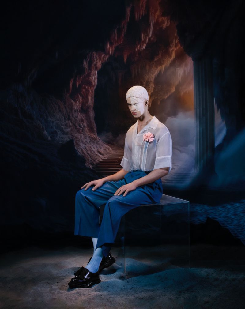 A male model with painted white skin, wearing a semi-sheer white shirt and blue pants, sits on a clear stool in a dark, moody studio.