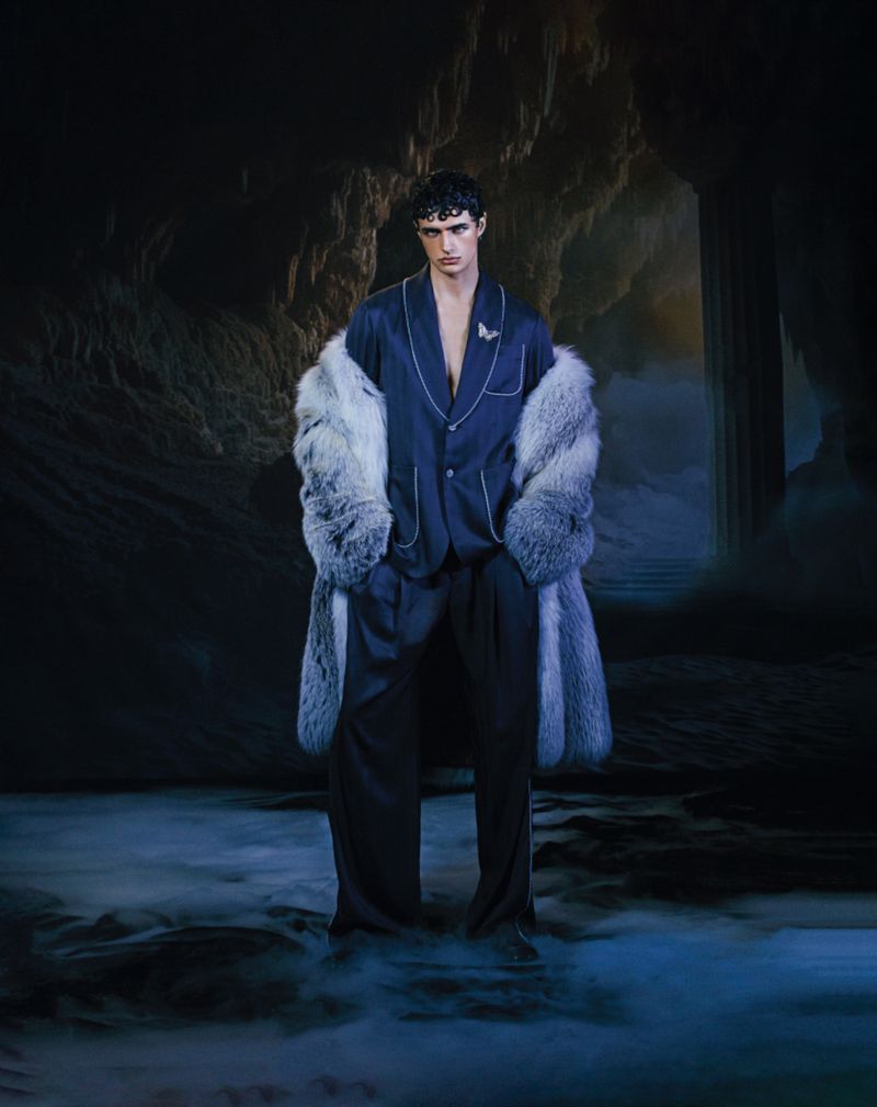 Man stands in navy lounge set with fur coat in a cave, creating a dramatic and striking fashion image with a dark, moody atmosphere.