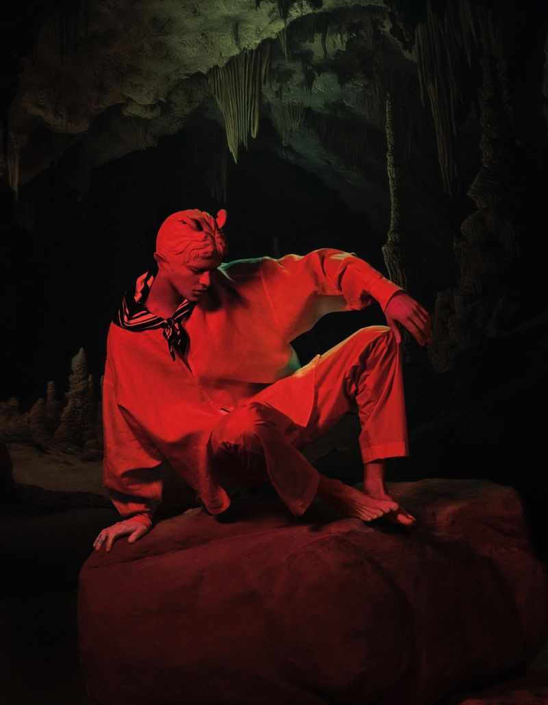 Man in sailor outfit sits on rock formation inside cave with stalactites, illuminated in red and green light.