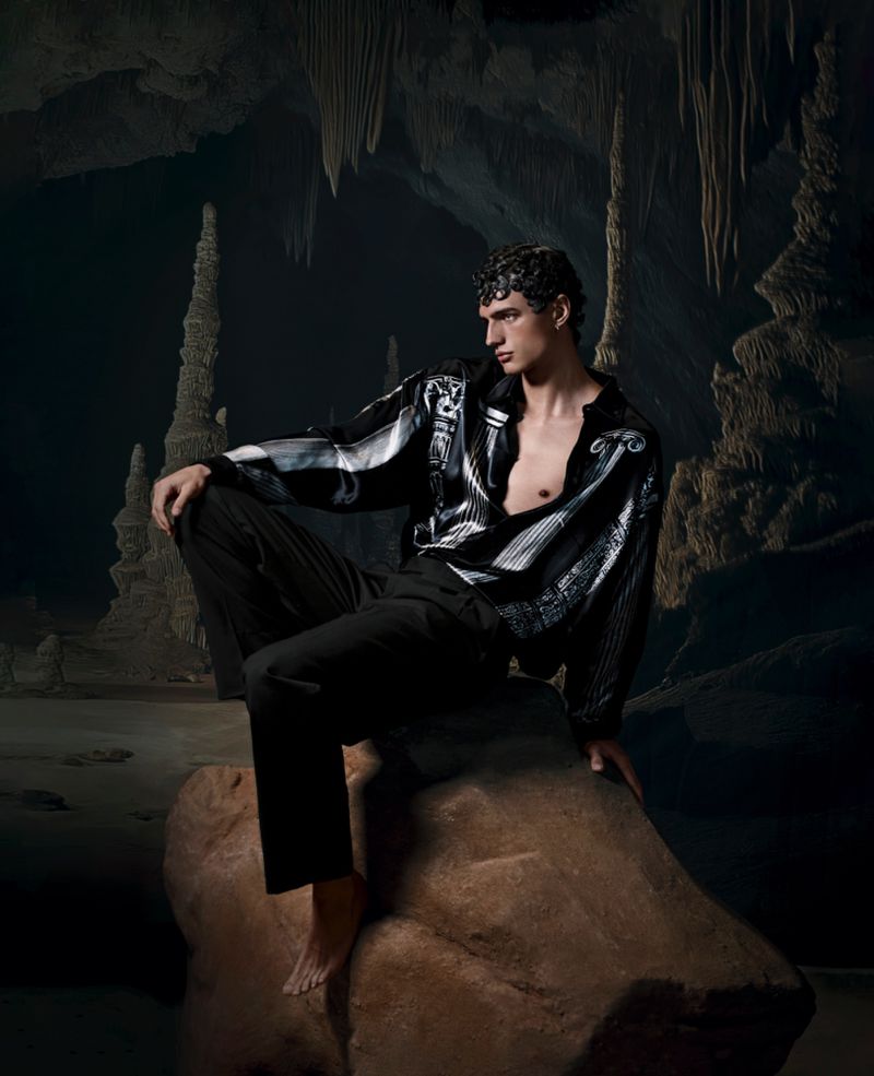 A male model poses on a rock formation inside a cave wearing a patterned shirt and black pants, illuminated by a spotlight.