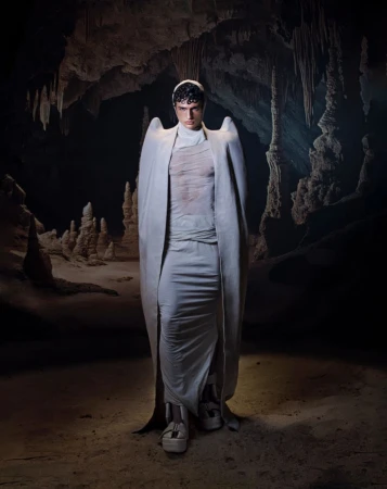 A pale, slender person in draped white garment stands in cave with hanging stalactites, platform shoes, and neutral facial expression.