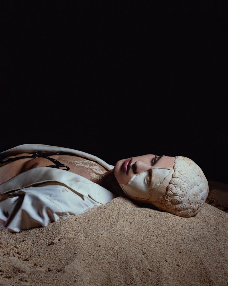 Surreal portrait of a person lying in sand with a partial sculpture mask, wearing a white shirt, against a dark, shadowy background.