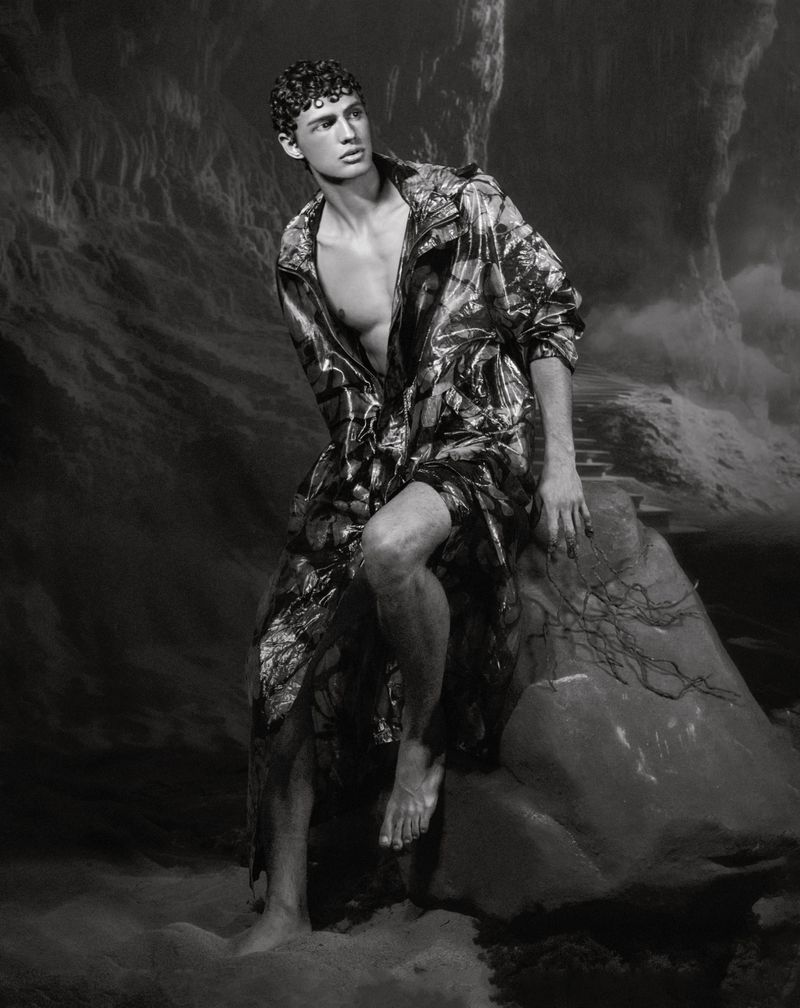 Monochrome portrait of a slender adult male with short curly hair, posing in an unbuttoned jacket while seated on a large rock.