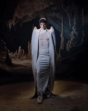 Model wearing sculptural cream cape and draped gown standing inside a dark limestone cave with stalagmites