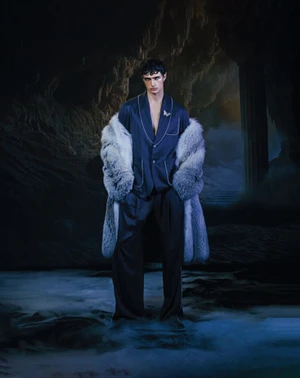 Model wearing a dark silk pajama-style jacket and fur coat standing in a cavernous rocky setting with dramatic lighting.