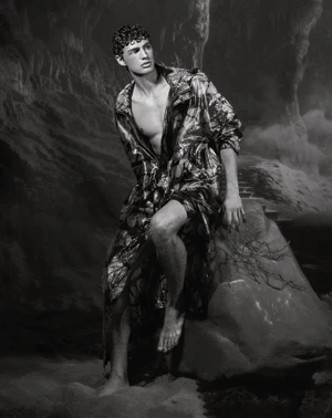 Monochrome fashion editorial featuring male model Ondrej Mokos seated on a stone in a cave setting wearing a patterned coat.