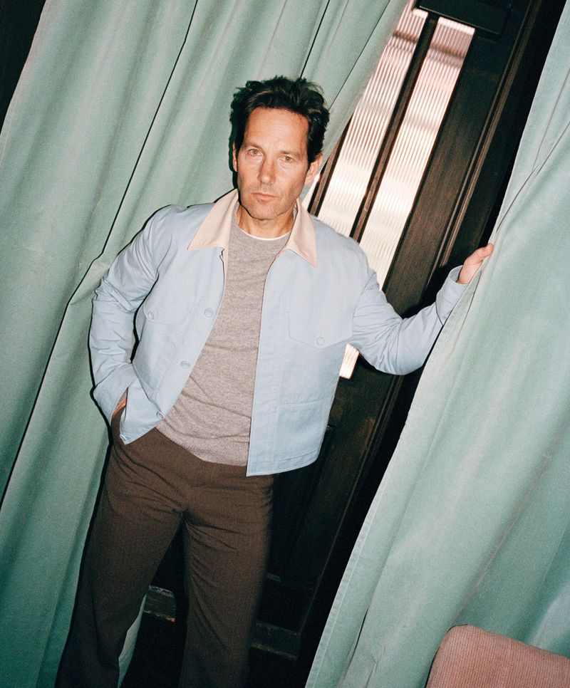 Portrait of Paul Rudd leaning against a window with light blue curtains, wearing a light blue jacket and brown trousers.