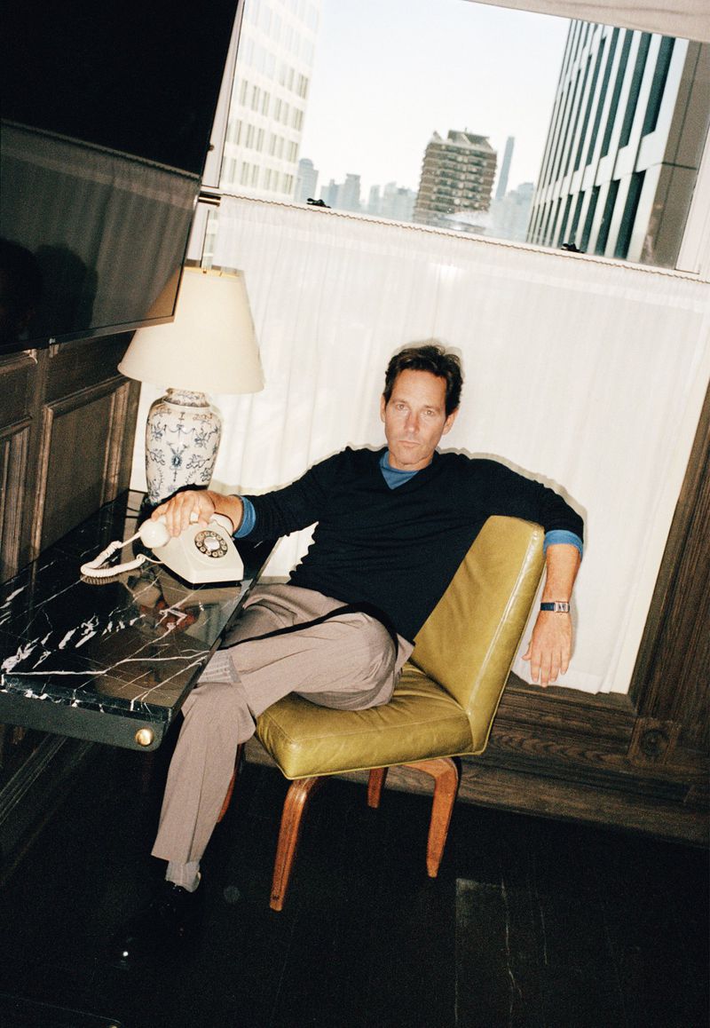 An adult male sits on a green chair indoors next to a marble-topped table with a vintage telephone and a view of city buildings in the background.
