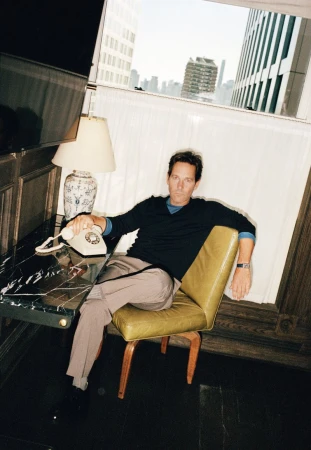 An adult male sits on a green chair indoors next to a marble-topped table with a vintage telephone and a view of city buildings in the background.