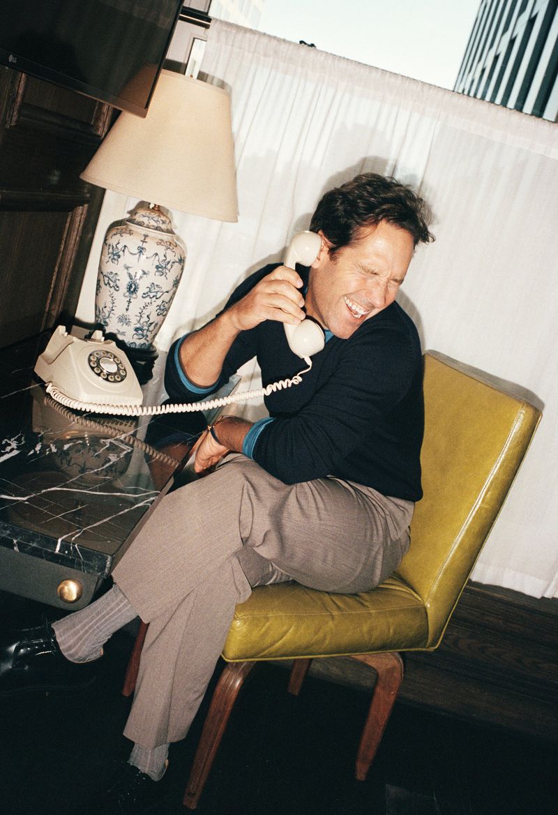 Adult man smiles while on a vintage phone in an indoor setting with a lamp, desk, and architectural window in the background.