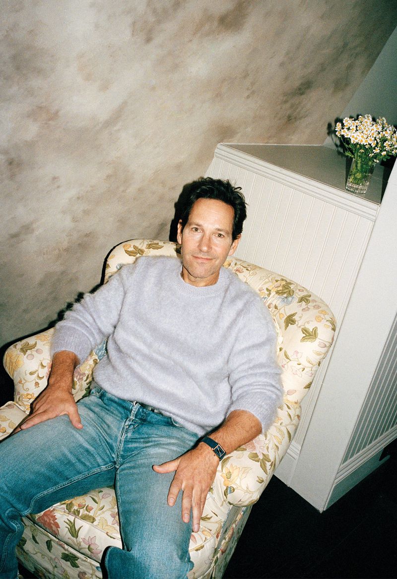 Indoor portrait of adult male sitting in a floral armchair with blue jeans and a light purple sweater in a house setting.