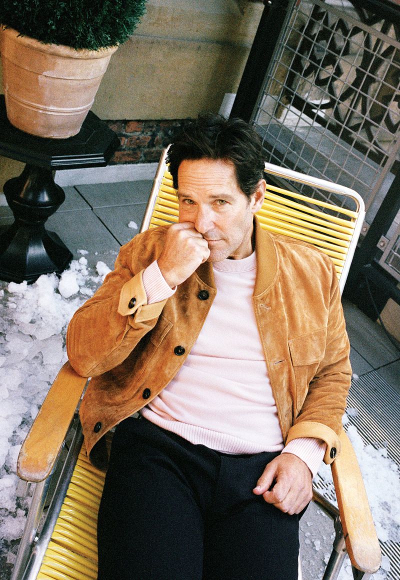 Portrait of actor Paul Rudd sitting on a yellow chair outside with a potted plant and metal grate in the background, wearing a tan jacket.