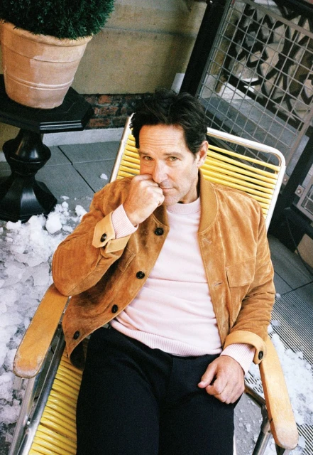 Portrait of actor Paul Rudd sitting on a yellow chair outside with a potted plant and metal grate in the background, wearing a tan jacket.