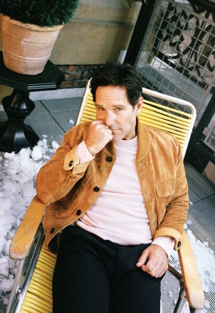 Portrait of actor Paul Rudd sitting on a yellow chair outside with a potted plant and metal grate in the background, wearing a tan jacket.