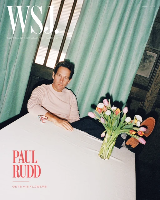 Paul Rudd sits at a white table beside a vase of colorful tulips. He is wearing a pink shirt. Teal curtain in background.