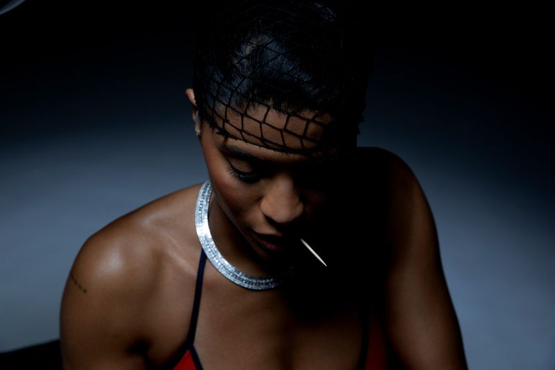 Close-up portrait of a woman with short dark hair, wearing a diamond necklace, against a dark gray background in low-key lighting.