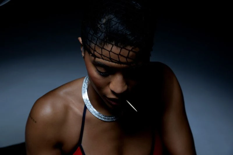 Close-up portrait of a woman with short dark hair, wearing a diamond necklace, against a dark gray background in low-key lighting.