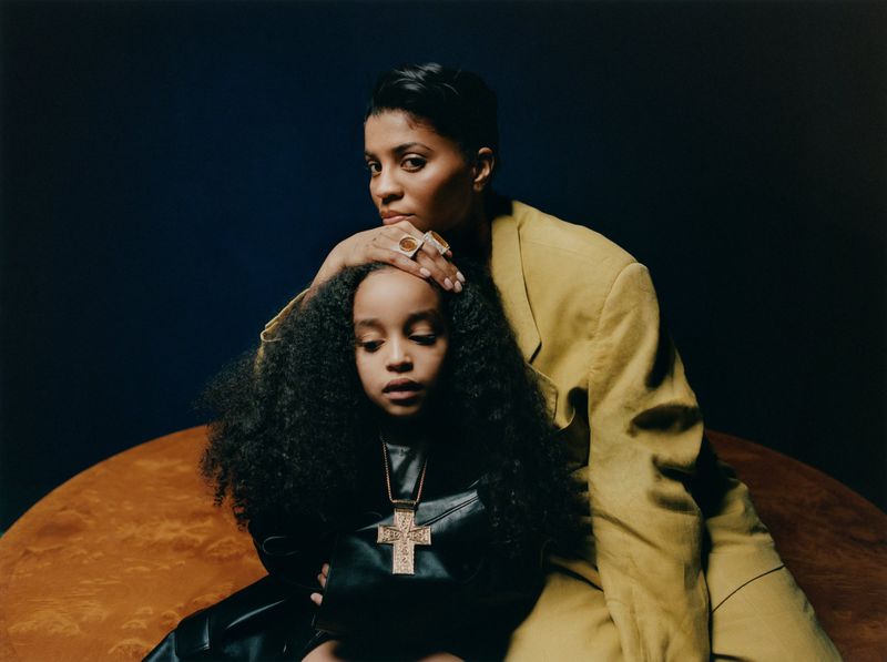 A woman in a mustard-colored blazer places her hand on the head of a child with dark curly hair wearing a black leather outfit and gold cross.