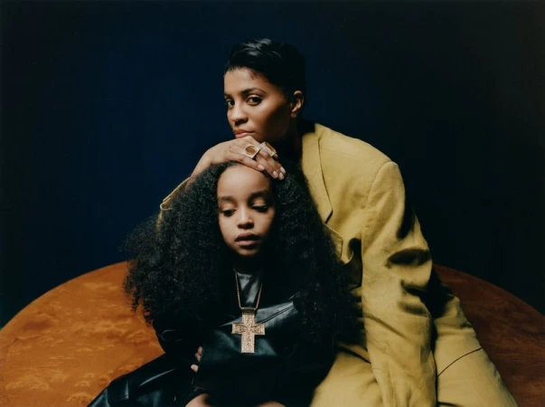 A woman in a mustard-colored blazer places her hand on the head of a child with dark curly hair wearing a black leather outfit and gold cross.