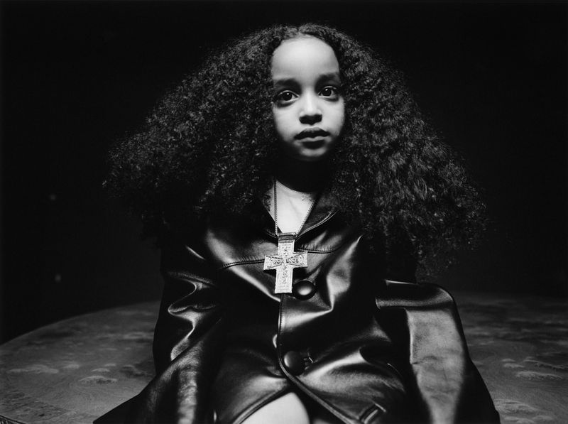 Monochrome portrait of a young African American girl in a leather jacket and cross pendant, set against a dark background.
