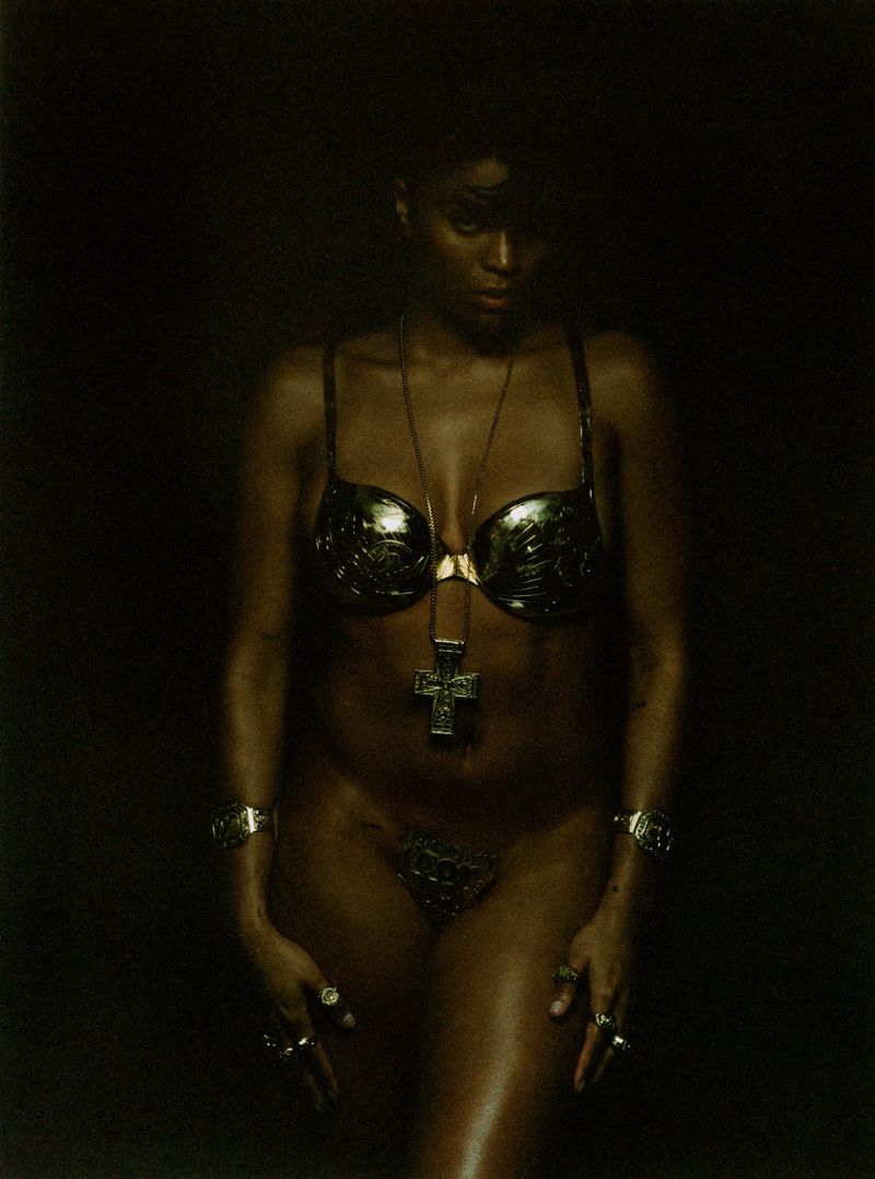Woman with dark skin in gold bra and bottom, cross pendant necklace, and jewelry on dark background, looking down.