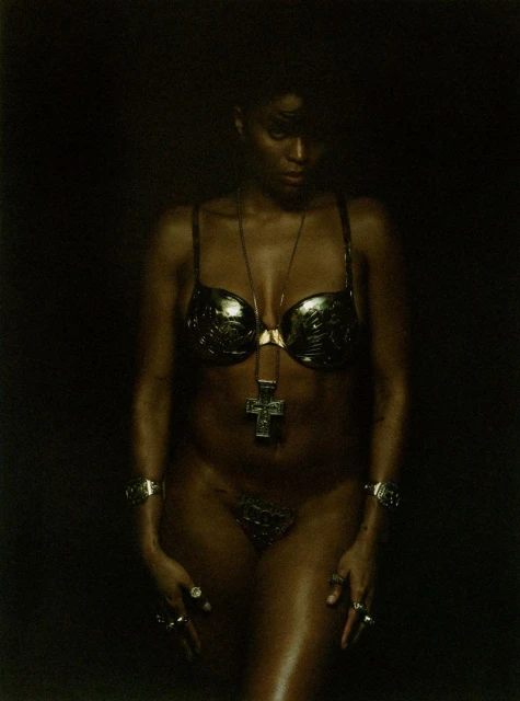 Woman with dark skin in gold bra and bottom, cross pendant necklace, and jewelry on dark background, looking down.