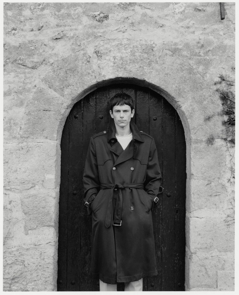 Monochrome portrait of a man in a trench coat, standing in front of an arched stone doorway, creating a classic and timeless image.