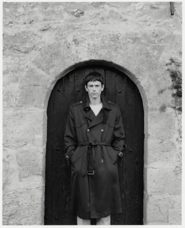 Monochrome portrait of a man in a trench coat, standing in front of an arched stone doorway, creating a classic and timeless image.