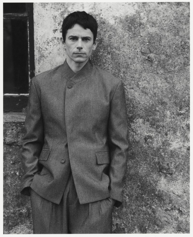 Monochrome portrait of a slender man wearing a tailored suit leaning against a textured wall near a window, creating a contemplative mood.