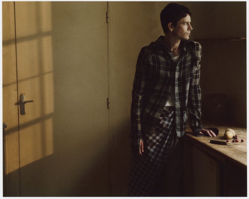 Man in a plaid shirt and pants leans against a wooden table in a dimly lit room with a window casting shadows on the door.