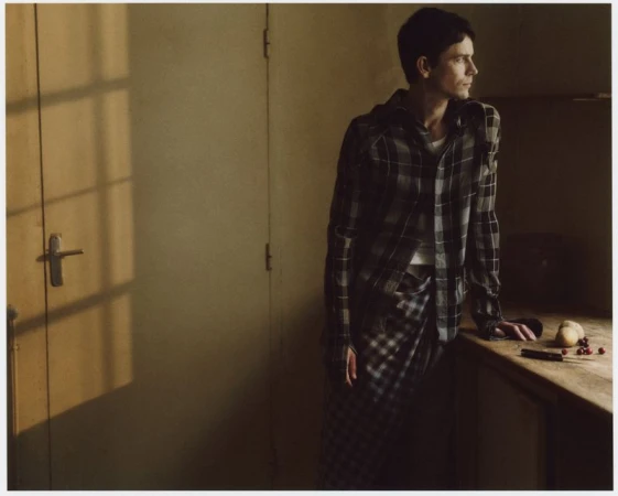 Man in a plaid shirt and pants leans against a wooden table in a dimly lit room with a window casting shadows on the door.