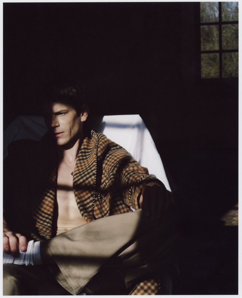 Indoor portrait of an adult male with brown hair seated in dappled lighting, wearing a tan and brown plaid jacket and khaki pants.