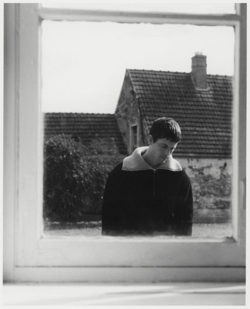 Black and white portrait of an adult male gazing downward, framed by a white window, with a stone house visible in the background.