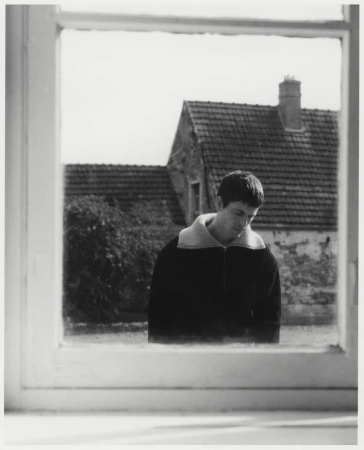 Black and white portrait of an adult male gazing downward, framed by a white window, with a stone house visible in the background.