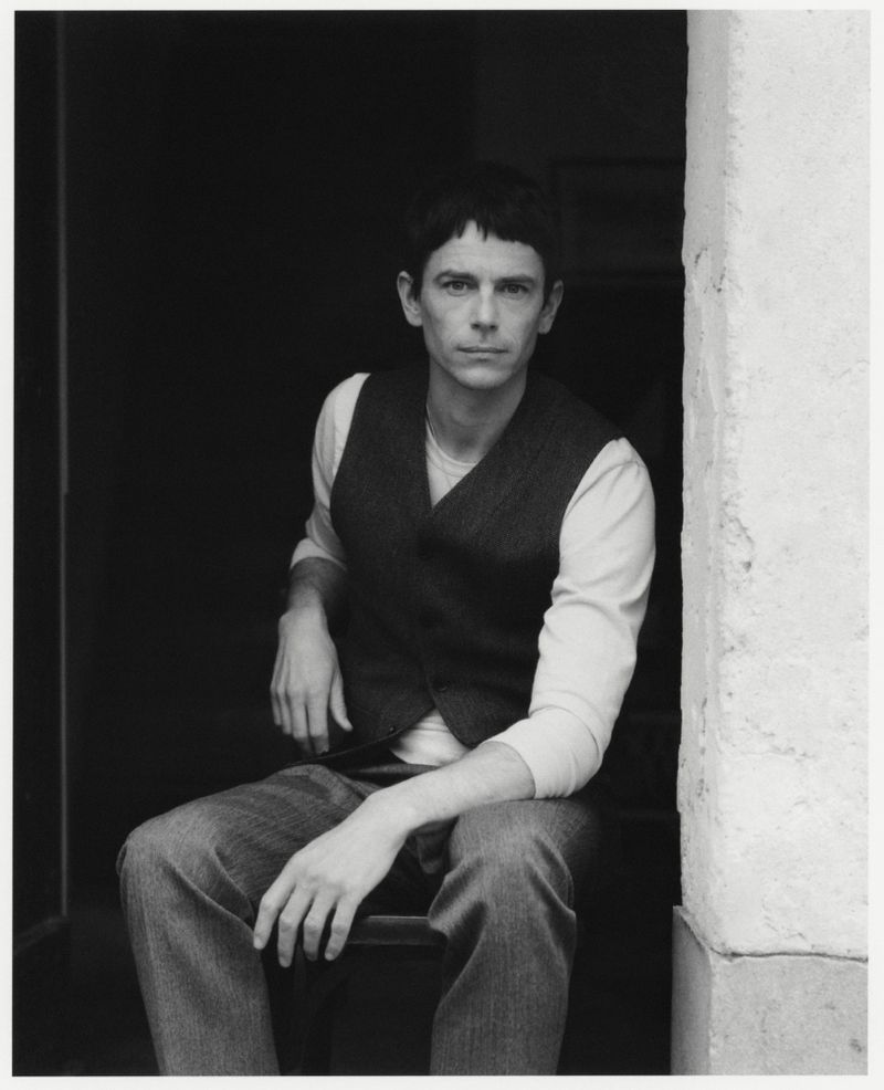 Portrait of a man with dark hair wearing a vest, long-sleeved shirt and jeans, seated in an indoor environment with natural light.