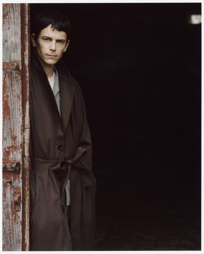 Portrait of a man with dark hair in a brown coat standing in a doorway with aged wood, captured with natural lighting.