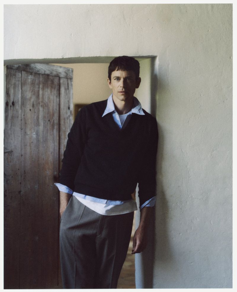 Indoor portrait of adult male leaning against a white wall next to a rustic wooden door, wearing a dark sweater over a collared shirt.