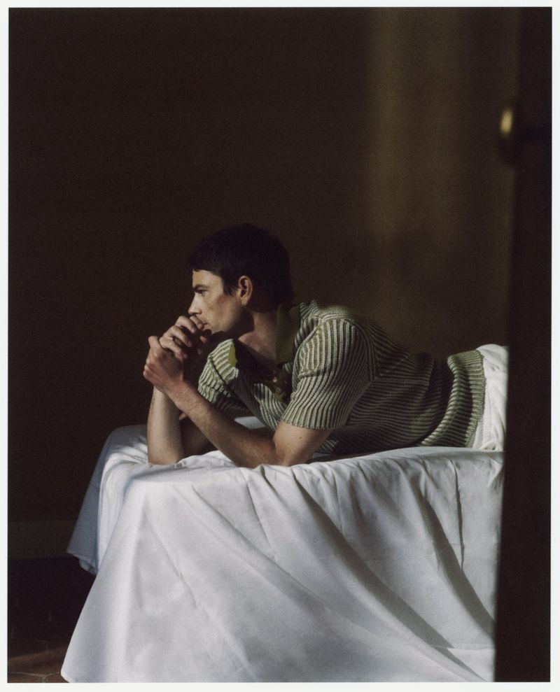 Man lying on a bed in a striped shirt with hands folded under his chin in a dimly lit room with neutral wall paint.