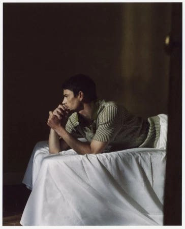 Man lying on a bed in a striped shirt with hands folded under his chin in a dimly lit room with neutral wall paint.