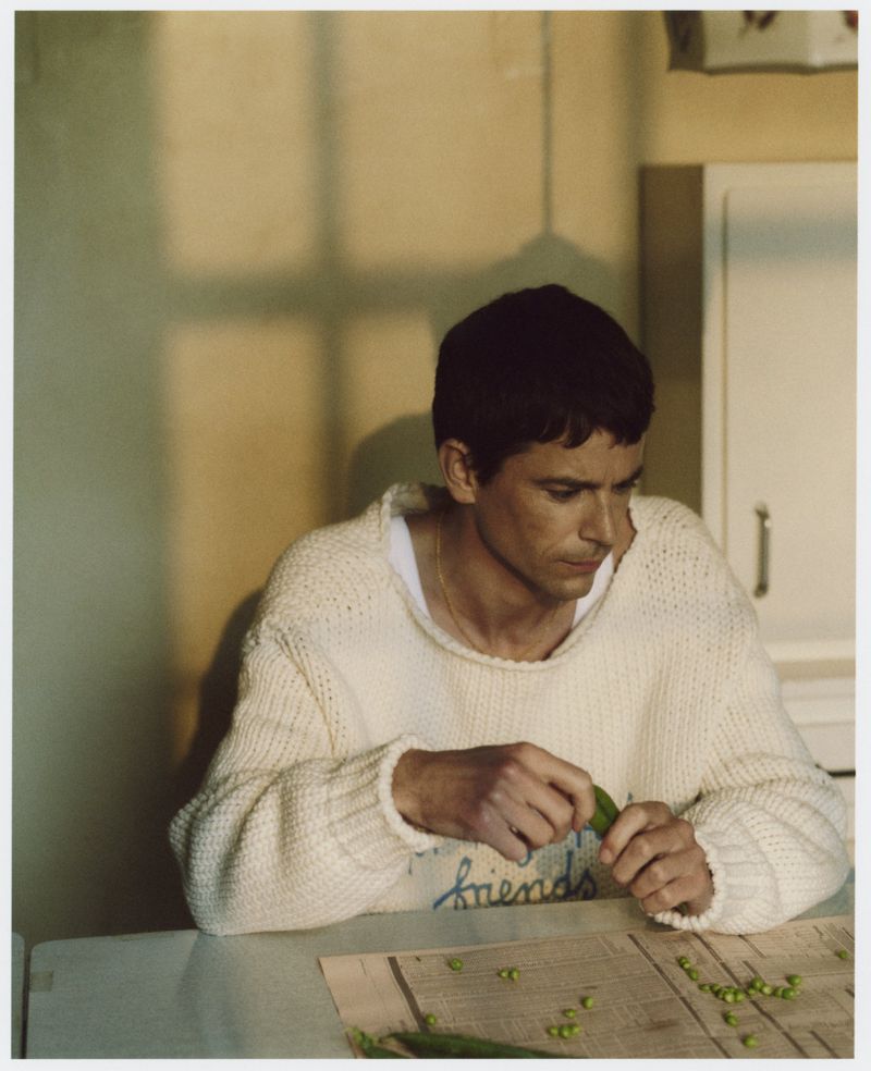Young man in a white sweater sits at a table in a kitchen peeling peas, with a newspaper and a window behind him.