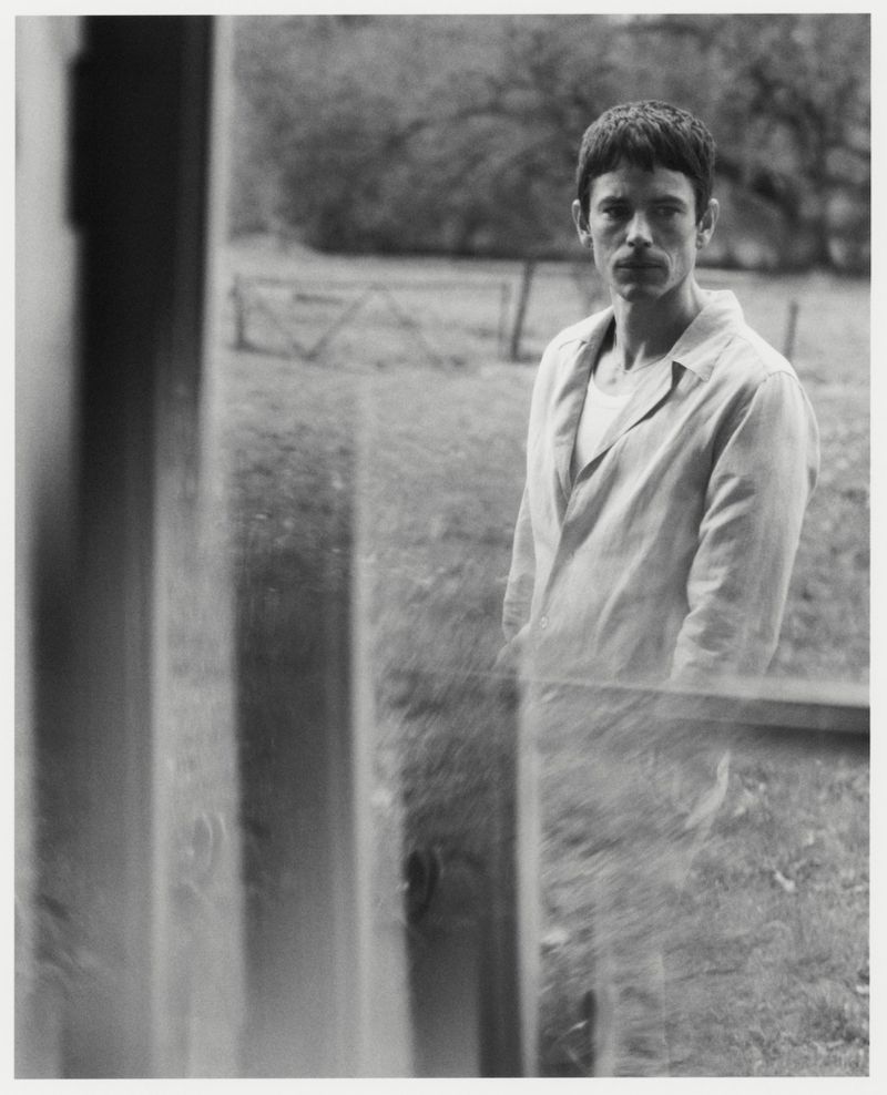 Black and white portrait of a man in an open collared shirt standing outdoors near a rustic fence.