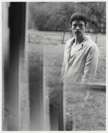 Black and white portrait of a man in an open collared shirt standing outdoors near a rustic fence.